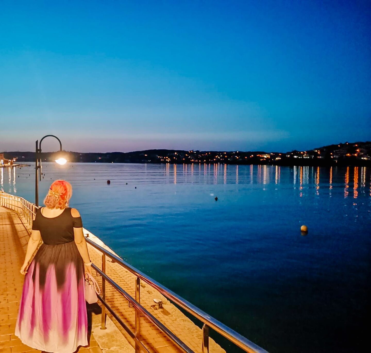 A woman with pink hair is looking out across the water at Cales Fonts, Es Castell