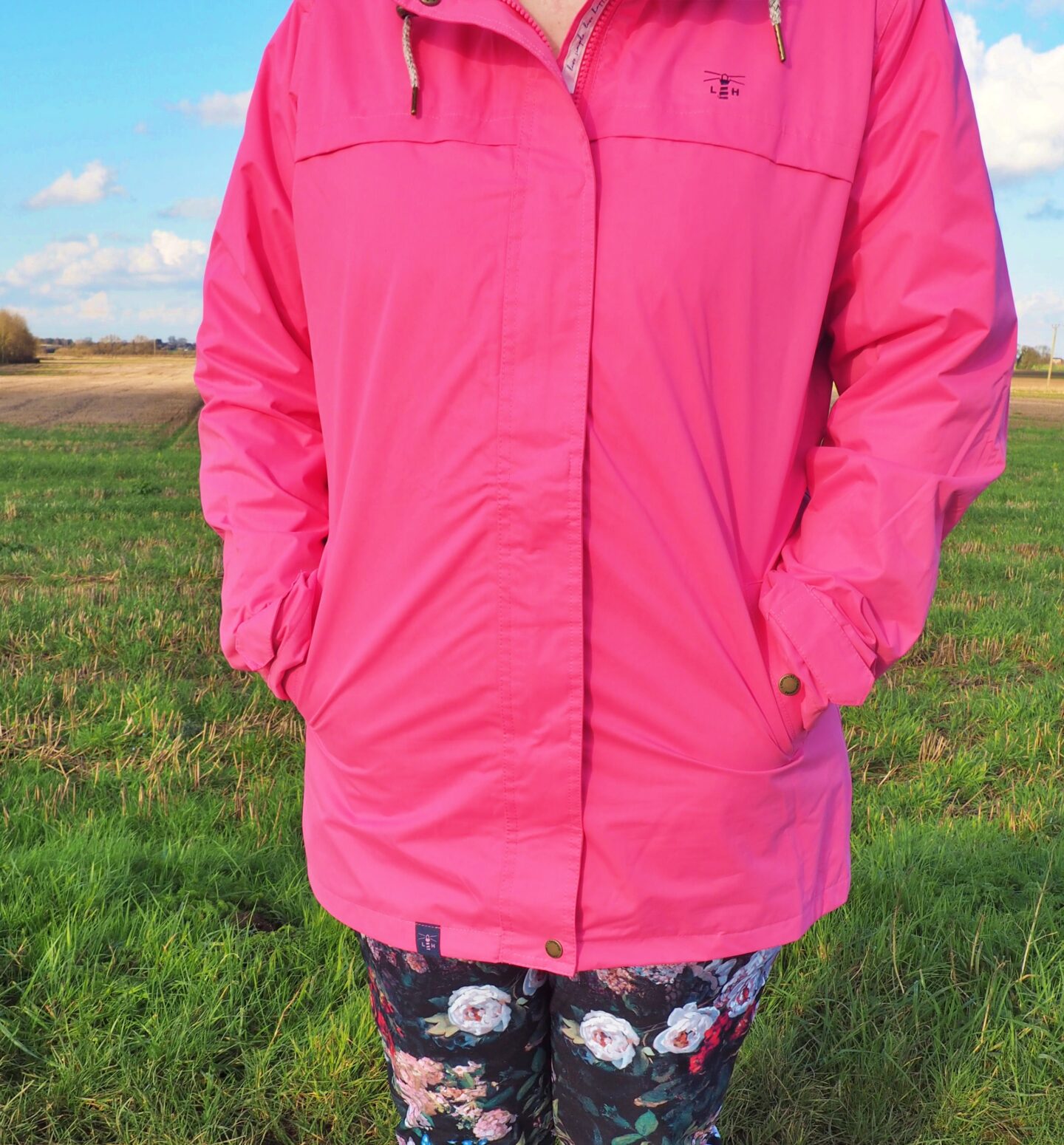 Image shows my shoulders down wearing a pink jacket. My hands are inside the pockets. I am stood on the edge of a field.