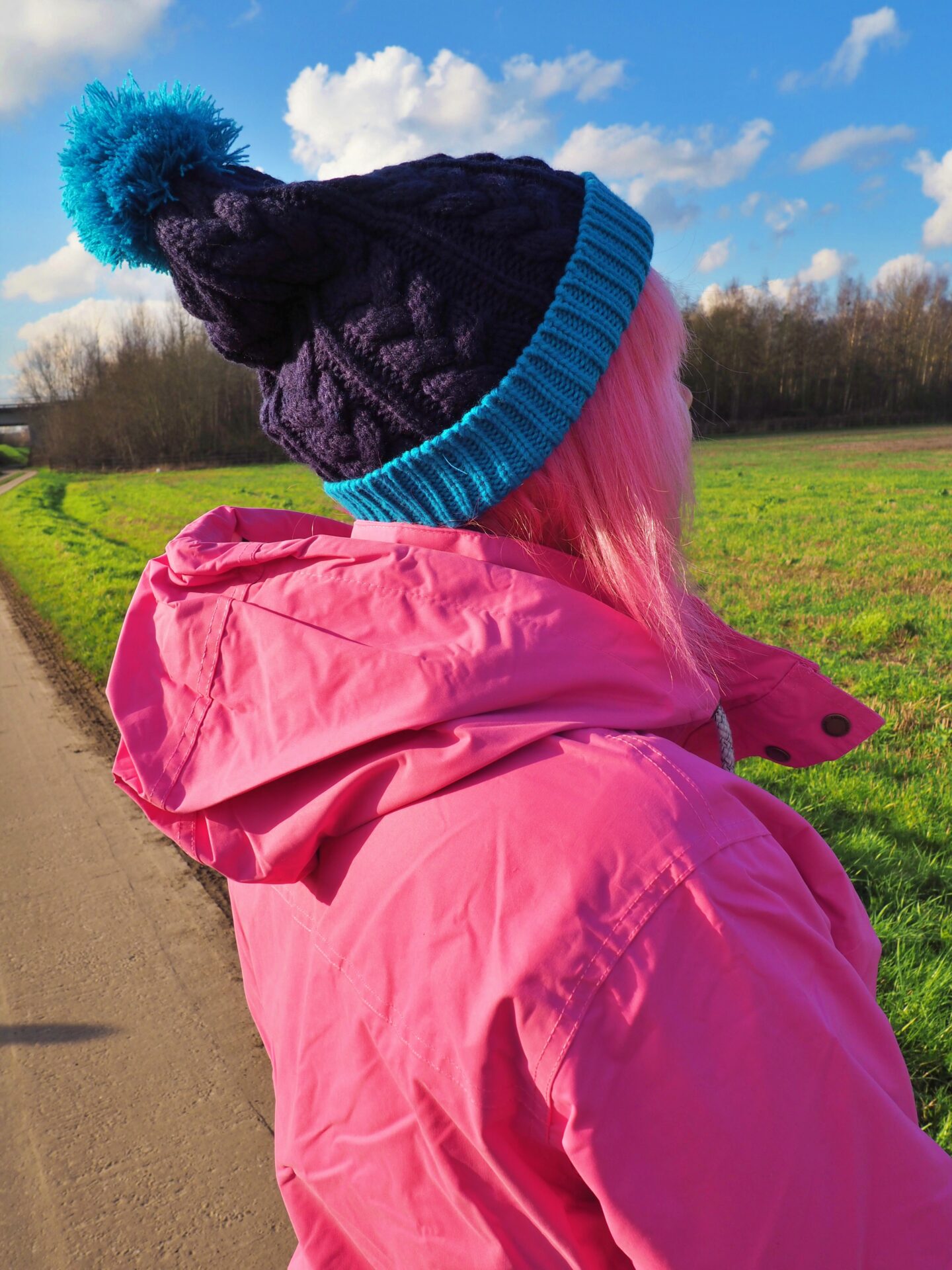 Image shows a woman with pink hair, wearing a pink jacket and a teal and navy coloured bobble hat. The angle the photo is taken from is from the right hand side and slightly behind me.