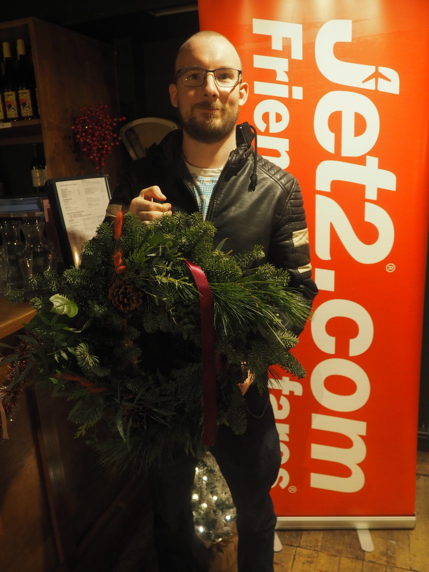 Tom, my husband, is stood in front of a red Jet2 banner. He is smiling and holding up the wreath he made during the event.