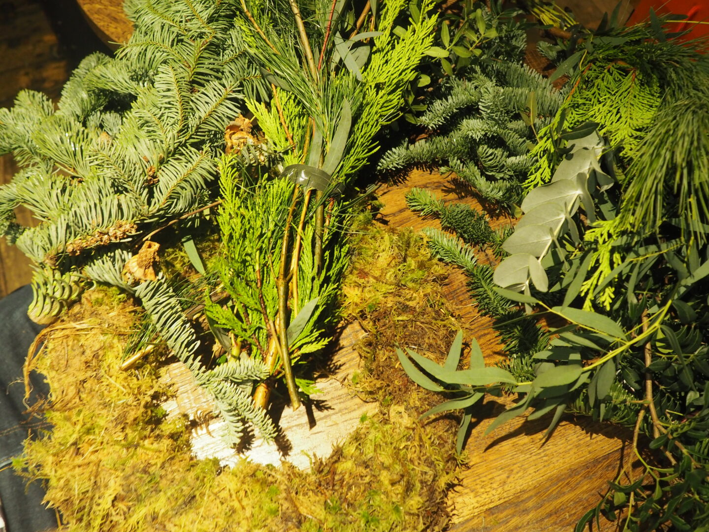 A table with a moss wreath wheel, around it are different pieces of cut offs and foliage. 